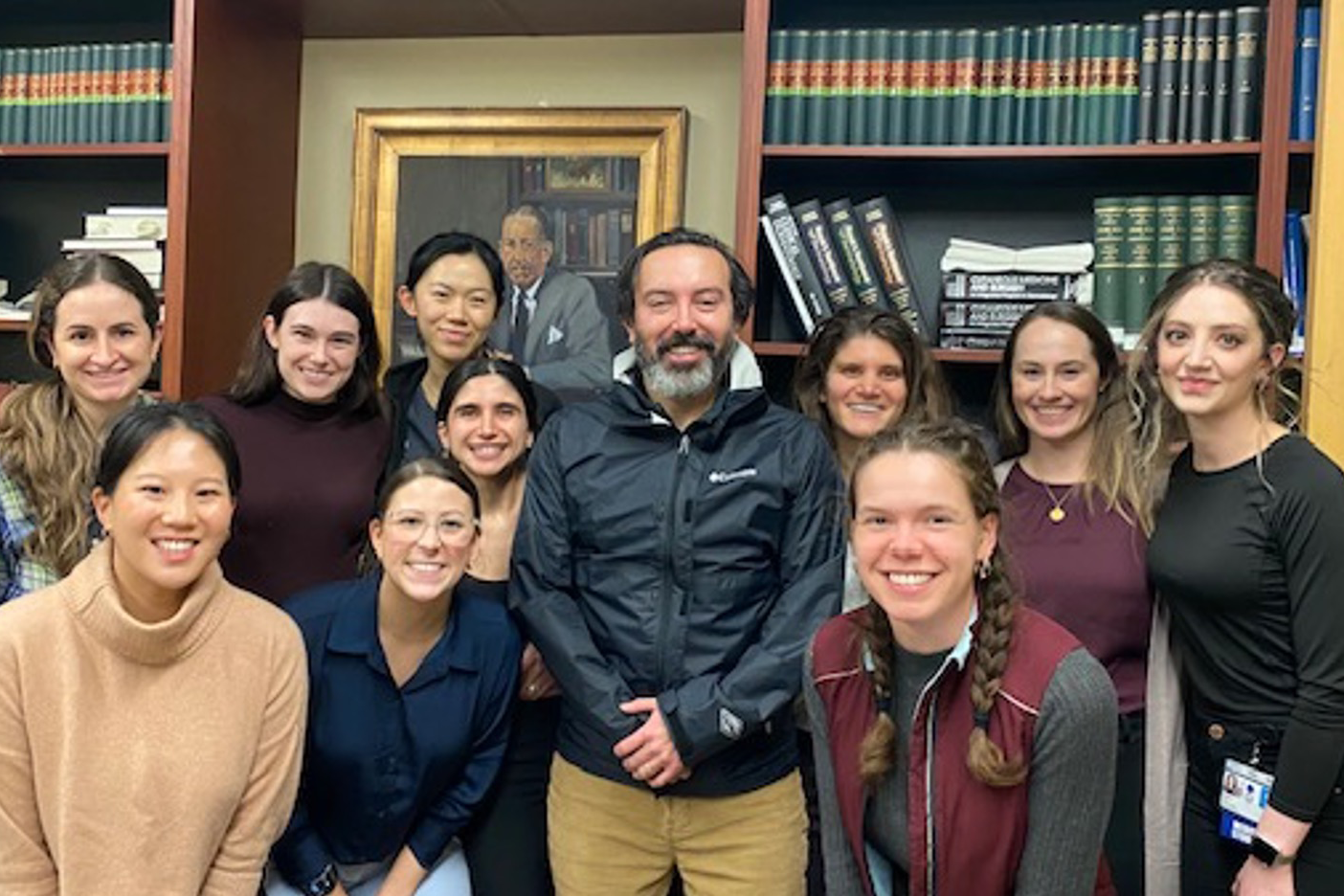Photo of Dr. Alex Ortega in a dark blue windbreaker jacket, in center, with a group of students around him in a semi-circle, against a background of books and a painting with a gilded frame.