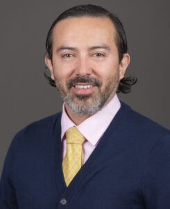 Headshot of Dr. Alex Ortega, professor and interim chair of Dermatology at Oregon Health & Science University, smiling in a dark blue jacket and pale-yellow shirt against a dark grey background.