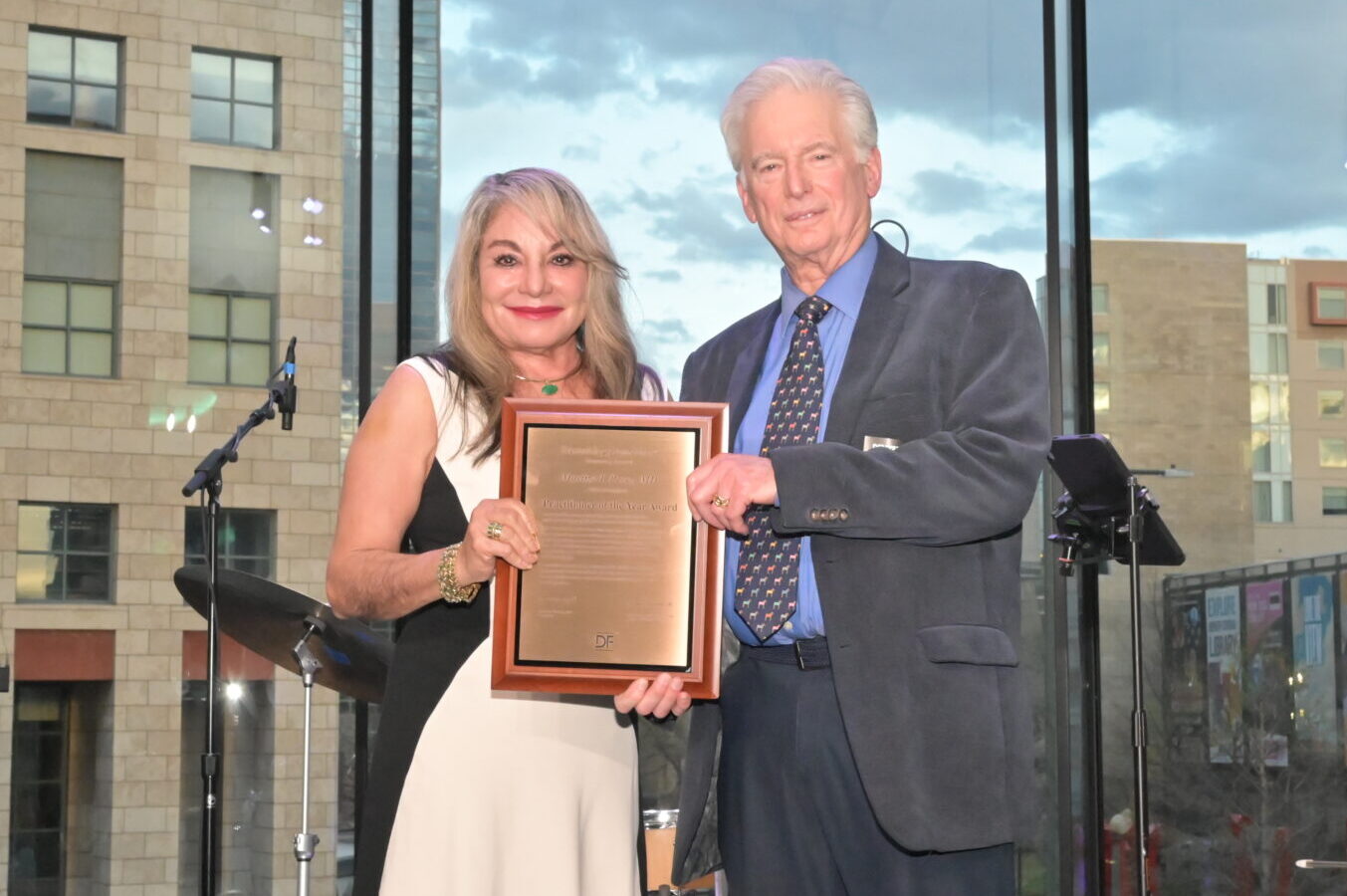 Female doctor wearing a dress in black and cream receives an award from a male doctor in a grey suit and light blue shirt.