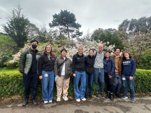 Photo of Dr. Tiffany Scharschmidt and eight members of her lab standing against a backdrop of hedges and trees.