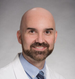 Headshot of Dr. Cory Simpson in a lab coat, wearing a pale blue shirt and a blue striped tie.