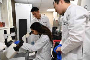 Two men standing and one woman seated at a microscope, wearing white lab coats, in a laboratory.