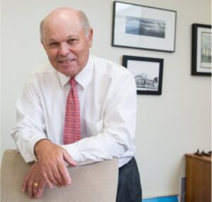 Man in white shirt and red tie in an office leans forward and rests his hands on a beige-colored upholstered chair.