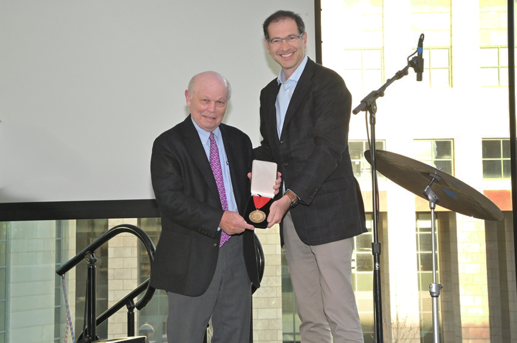 Two men in dark suits on a stage. The man on the left receives an award from the man on the right.